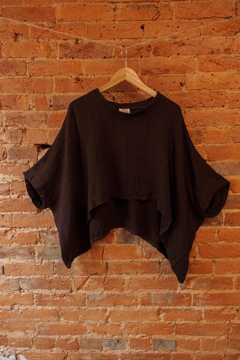 May include: A dark brown, loose-fitting, short-sleeved top with a rounded neckline. The top is hanging on a wooden hanger against a brick wall.