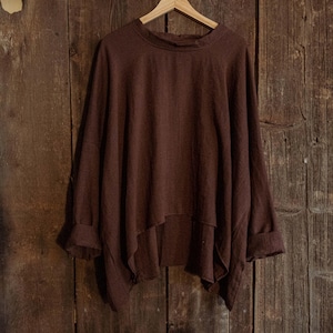Op de afbeelding: Een loszittende, langarmige blouse in bruin, hangend aan een houten hanger tegen een rustieke houten achtergrond.