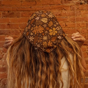 May include: A person with long brown hair is wearing a brown bandana with a floral pattern. The bandana has yellow, orange, and white flowers and leaves. The person is wearing a white tank top.