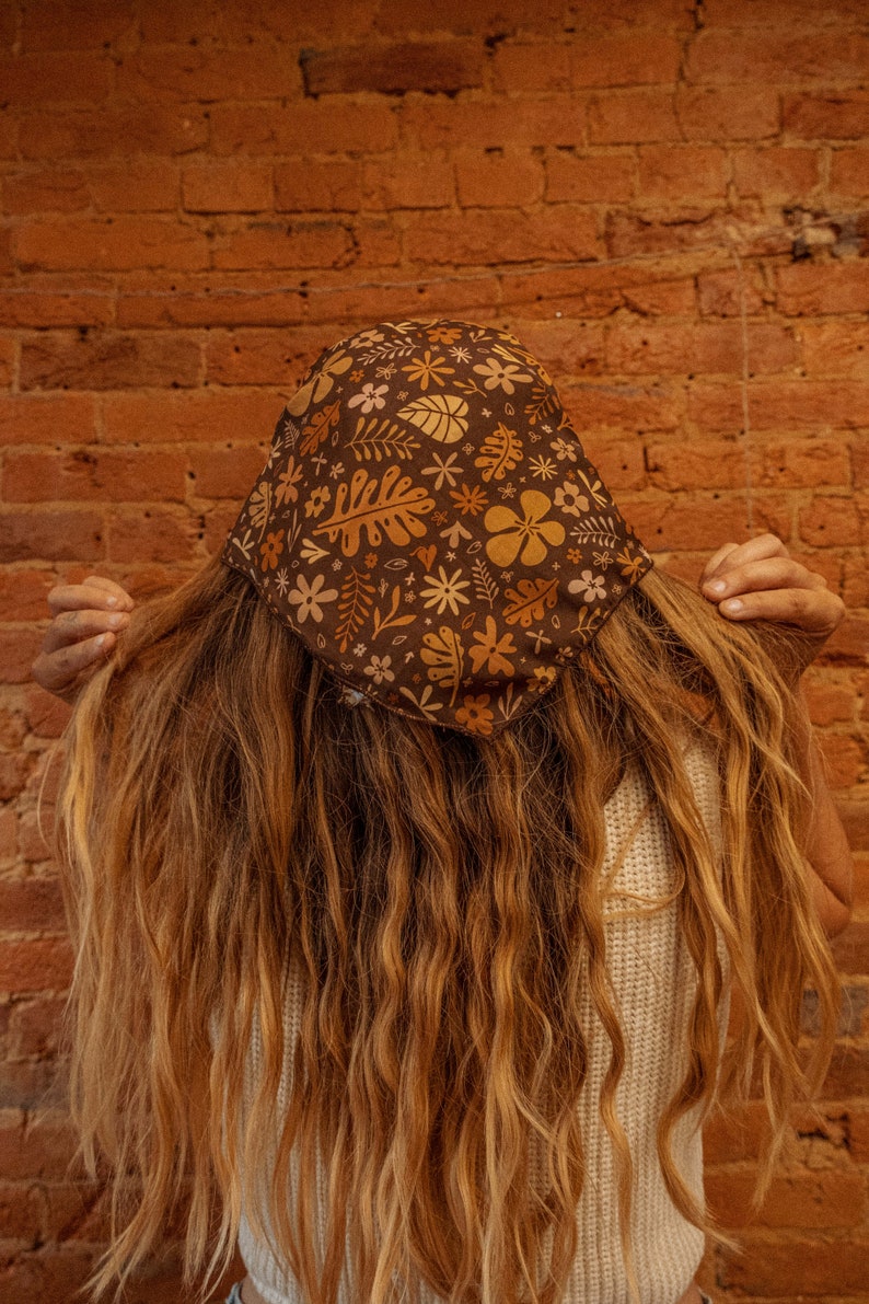 May include: A person with long brown hair is wearing a brown bandana with a floral pattern. The bandana has yellow, orange, and white flowers and leaves. The person is wearing a white tank top.