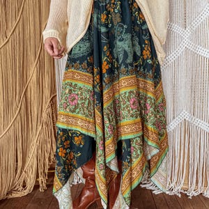 May include: A long, flowing skirt with a dark teal background and a colorful floral and paisley pattern. The skirt features bands of green, orange, and pink, and is paired with brown boots and a cream-colored sweater.