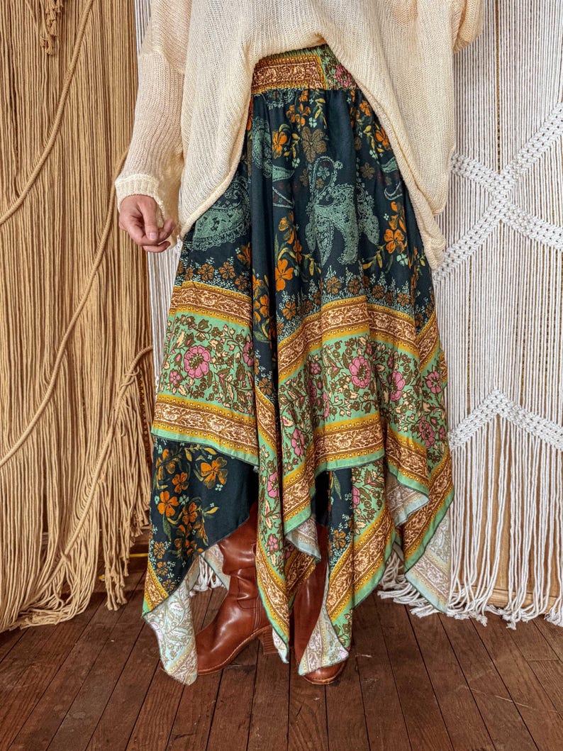 May include: A long, flowing skirt with a dark teal background and a colorful floral and paisley pattern. The skirt features bands of green, orange, and pink, and is paired with brown boots and a cream-colored sweater.