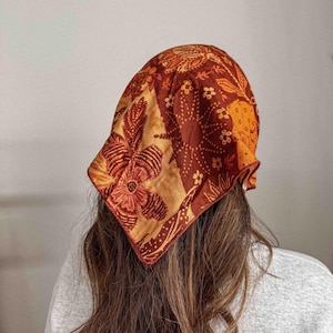 Puede incluir: Un pa&ntilde;uelo con estampado en tonos naranja, marr&oacute;n y crema, con dise&ntilde;os florales y de hojas. El pa&ntilde;uelo es cuadrado y est&aacute; atado alrededor de la cabeza. La tela parece suave y ligera.