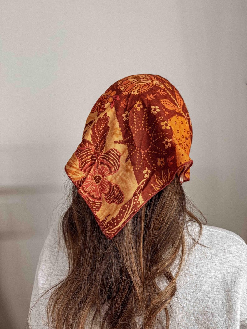 Puede incluir: Un pa&ntilde;uelo con estampado en tonos naranja, marr&oacute;n y crema, con dise&ntilde;os florales y de hojas. El pa&ntilde;uelo es cuadrado y est&aacute; atado alrededor de la cabeza. La tela parece suave y ligera.