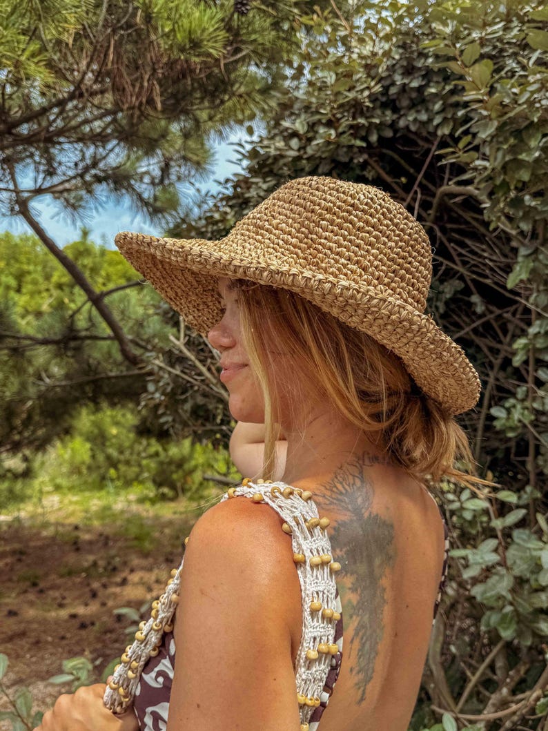 Pu&ograve; includere: Un cappello da sole in paglia intrecciata con una tesa larga. Il cappello &egrave; di colore marrone naturale e ha un aspetto leggermente strutturato. Il cappello &egrave; indossato all'aperto, con uno sfondo sfocato di alberi e fogliame.