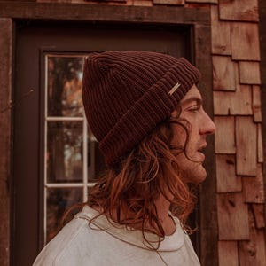 May include: A dark brown ribbed knit beanie with a small gold tag. The beanie is worn by a person with long, wavy auburn hair, wearing a white crew neck shirt. The background features a wooden door and a shingled wall.