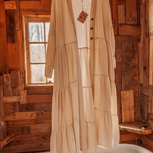 以下が含まれることがあります： ボタン留めの白い、ティアード、長袖の綿のカーディガン。カーディガンは、素朴なバスルームの環境で、木製ハンガーにかけられています。