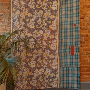 K&ouml;nnte beinhalten: Eine Vintage-Kantha-Steppdecke mit einem floralen Muster auf der einen Seite und einem karierten Muster auf der anderen Seite. Die Steppdecke h&auml;ngt an einer Backsteinwand.