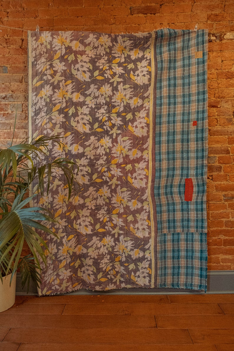K&ouml;nnte beinhalten: Eine Vintage-Kantha-Steppdecke mit einem floralen Muster auf der einen Seite und einem karierten Muster auf der anderen Seite. Die Steppdecke h&auml;ngt an einer Backsteinwand.