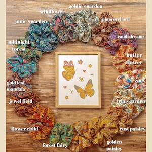 May include: A circle of colorful fabric scrunchies with floral patterns and names like "Goldie's Garden", "Wildflower", "Junie's Garden", "Midnight Forest", "Gold Leaf Mandala", "Jewel Field", "Flower Child", "Forest Fairy", "Honey Bee", "Golden Paisley", "Rust Paisley", "Lyla's Garden", "Zoev", "Butter Flower", "Candi Dreams", and "Plum Orchard". A framed picture of two butterflies with a floral background is in the center.