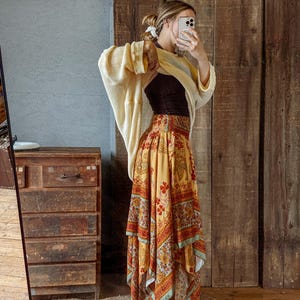 May include: A woman wearing a cream-colored cardigan over a black top and a long, flowing skirt. The skirt features a vibrant, patterned design in shades of yellow, red, and blue. The background includes a wooden door and a jute rug.