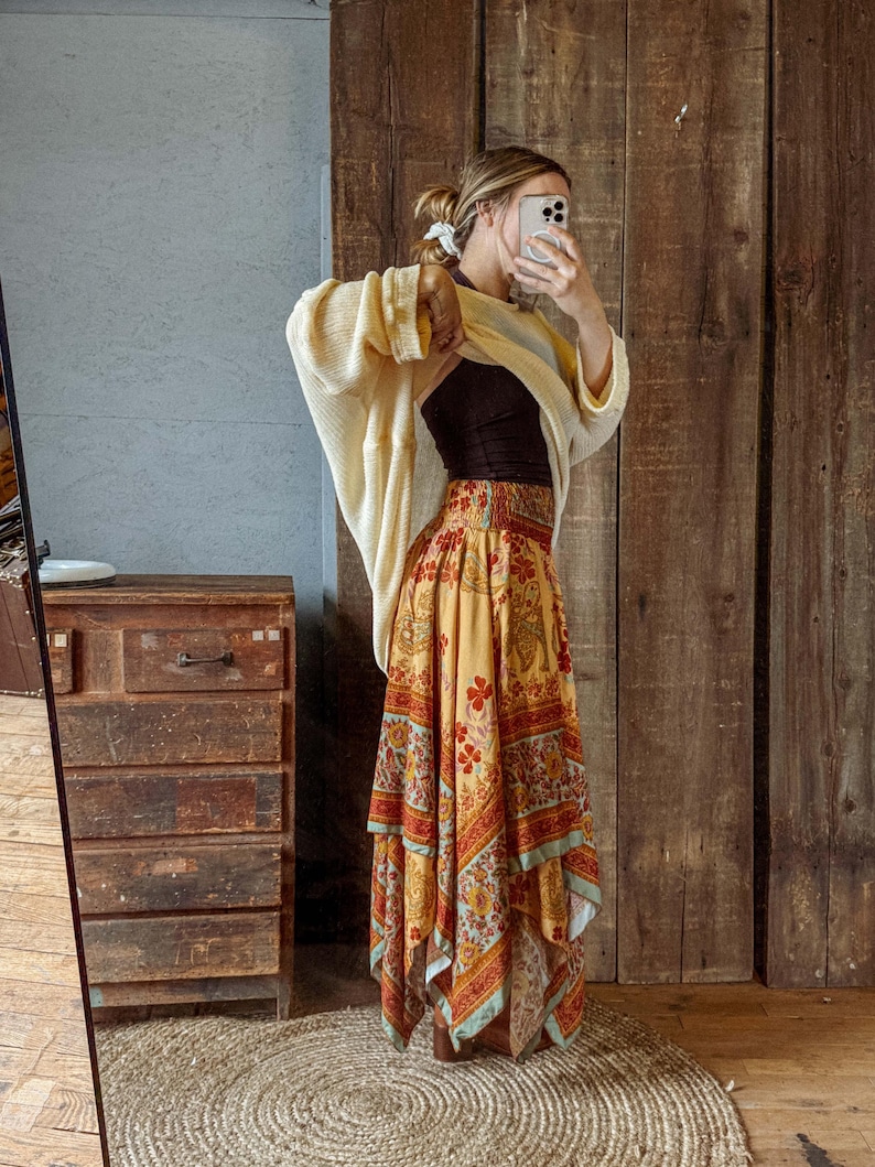 May include: A woman wearing a cream-colored cardigan over a black top and a long, flowing skirt. The skirt features a vibrant, patterned design in shades of yellow, red, and blue. The background includes a wooden door and a jute rug.