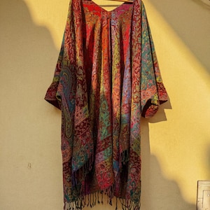 Könnte beinhalten: Ein farbenfrohes, gemustertes Tuch mit V-Ausschnitt, das an einem Holzbügel hängt. Das Tuch zeigt ein Paisley-Muster in Rot-, Blau-, Grün- und Lilatönen mit Fransen am unteren Rand. Das Kleidungsstück wird vor einer hellgelben Wand präsentiert.