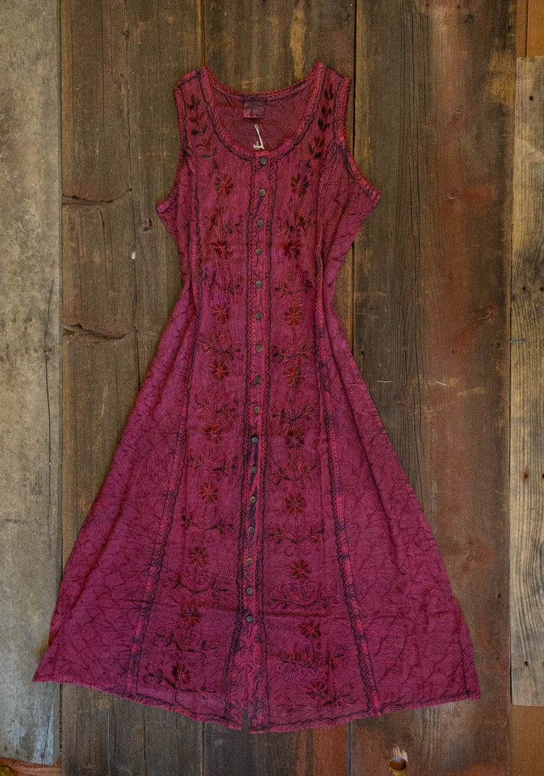 May include: A sleeveless, button-down dress in a deep magenta hue, adorned with intricate floral embroidery. The dress features a flared skirt and is displayed on a rustic wooden surface, showcasing its bohemian style.