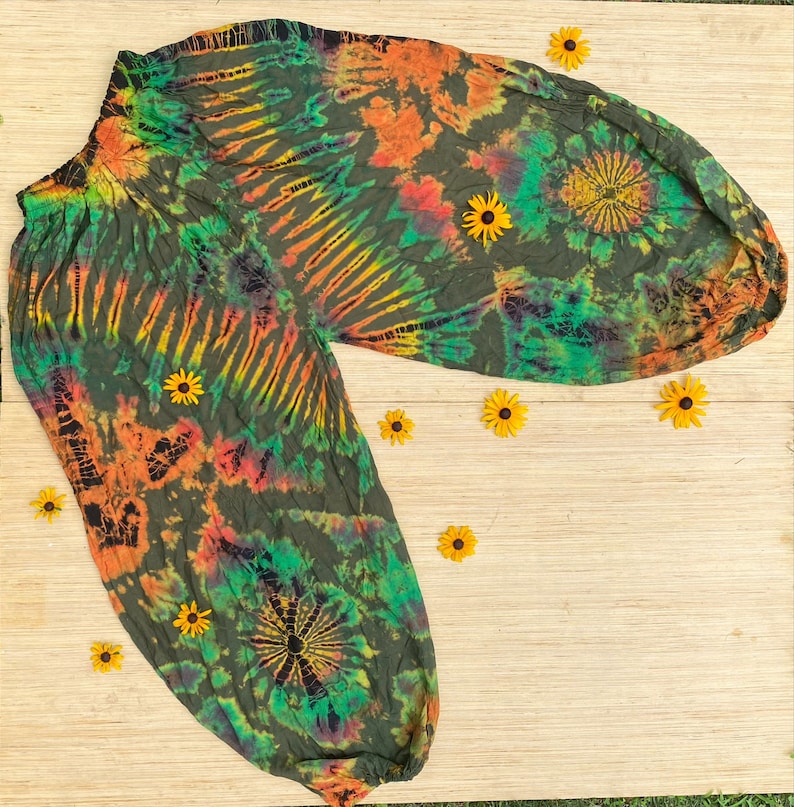 May include: Tie-dye trousers in green, orange, and yellow hues. The trousers feature an elasticated waistband and a loose fit. Yellow flowers are arranged around the garment on a light brown surface.