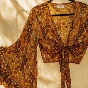 May include: A yellow, brown, and green floral print blouse with a tie front and long bell sleeves. The blouse has a V-neckline and a ruffled hem. The label reads "Gardenville Baby".