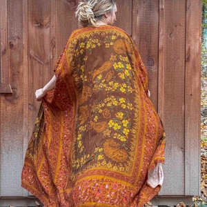 May include: A long, flowing kimono in shades of brown, orange, and yellow. The fabric features a floral and paisley design. The garment is worn outdoors, with a wooden wall in the background.