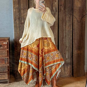May include: Cream-colored, loose-fitting sweater paired with a long, patterned skirt in shades of yellow, orange, and red. The skirt has a unique handkerchief hemline. Brown boots complete the outfit, creating a bohemian style.