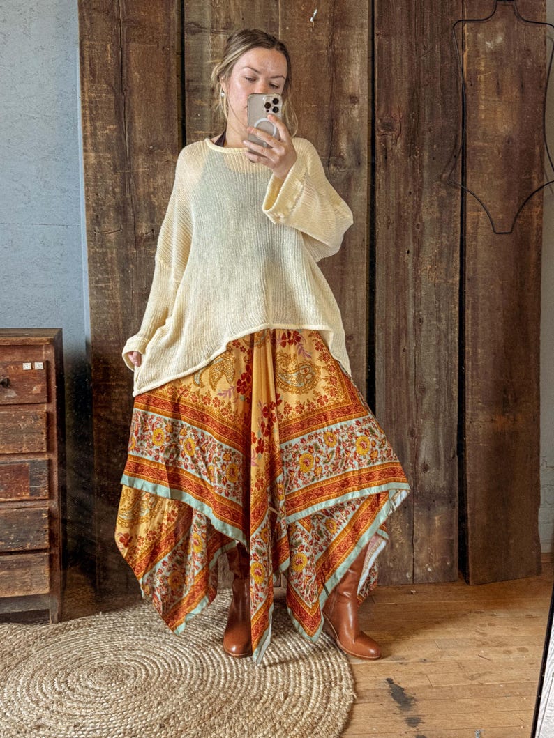 May include: Cream-colored, loose-fitting sweater paired with a long, patterned skirt in shades of yellow, orange, and red. The skirt has a unique handkerchief hemline. Brown boots complete the outfit, creating a bohemian style.