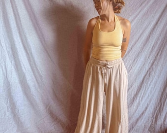 May include: A pale yellow halter top with a scoop neckline and a ruched bodice paired with loose-fitting, wide-leg beige trousers featuring an elasticized waistband with a drawstring tie, creating a soft and relaxed two-piece clothing set.