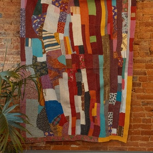 May include: A colorful patchwork quilt with a variety of fabrics and patterns, including stripes, florals, and geometric designs. The quilt is hanging on a brick wall.