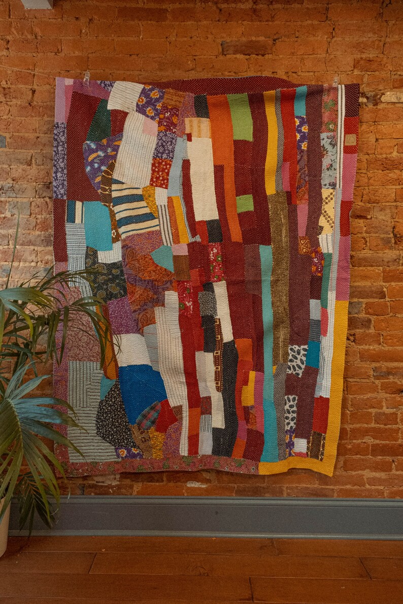 May include: A colorful patchwork quilt with a variety of fabrics and patterns, including stripes, florals, and geometric designs. The quilt is hanging on a brick wall.