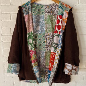 May include: A brown hooded jacket with a vibrant patchwork lining. The jacket is displayed on a wooden hanger against a white brick wall. The lining showcases floral and fruit patterns in multiple colours.
