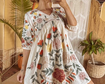 May include: A white, short-sleeved dress with a V-neck, adorned with colorful floral embroidery. The embroidery features red, yellow, and green flowers and leaves. The dress has a loose, flowing silhouette.