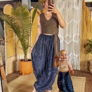 May include: A woman and a child are wearing matching tie-dye harem trousers. The woman is wearing a brown wrap top. The child is wearing a striped vest. The background includes a woven room divider, a macrame wall hanging, and a potted plant.