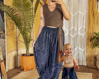 May include: A woman and a child wearing matching tie-dye harem pants. The woman is wearing a brown wrap top. The child is wearing a striped tank top. The background includes a woven room divider and a macrame wall hanging.