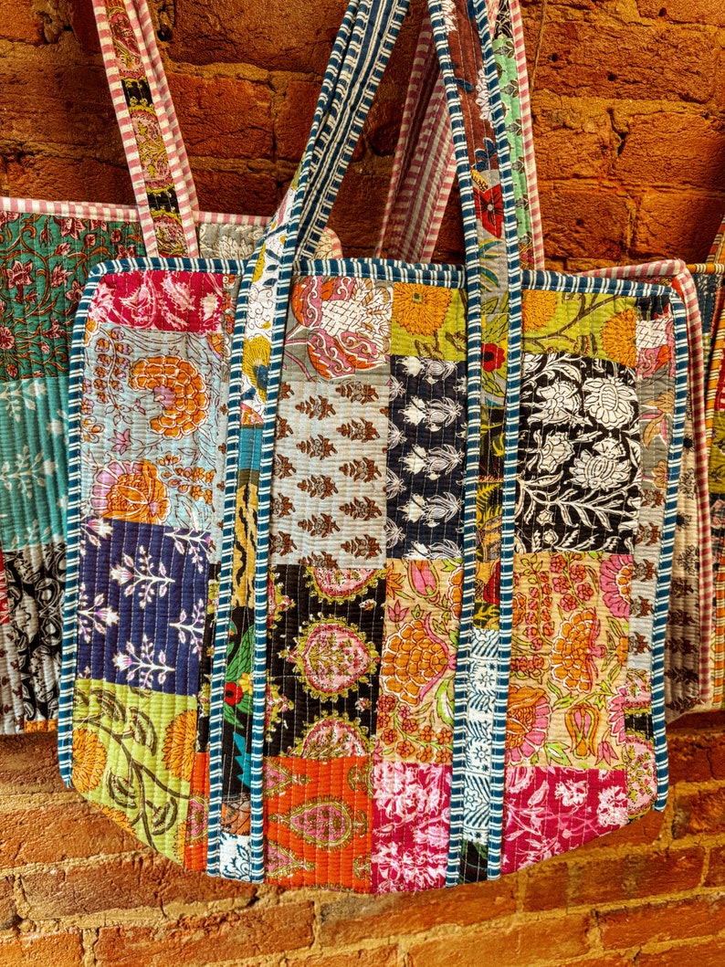 May include: Three colorful patchwork tote bags with blue and white striped straps. The bags are made of different fabrics with floral and geometric patterns.
