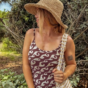 Pu&ograve; includere: Un cappello da sole in paglia intrecciata con tesa larga. &Egrave; abbinato a un abito marrone e bianco con motivo a onde e una borsa a tracolla color crema con perline. L'immagine &egrave; scattata all'aperto con uno sfondo verde naturale.