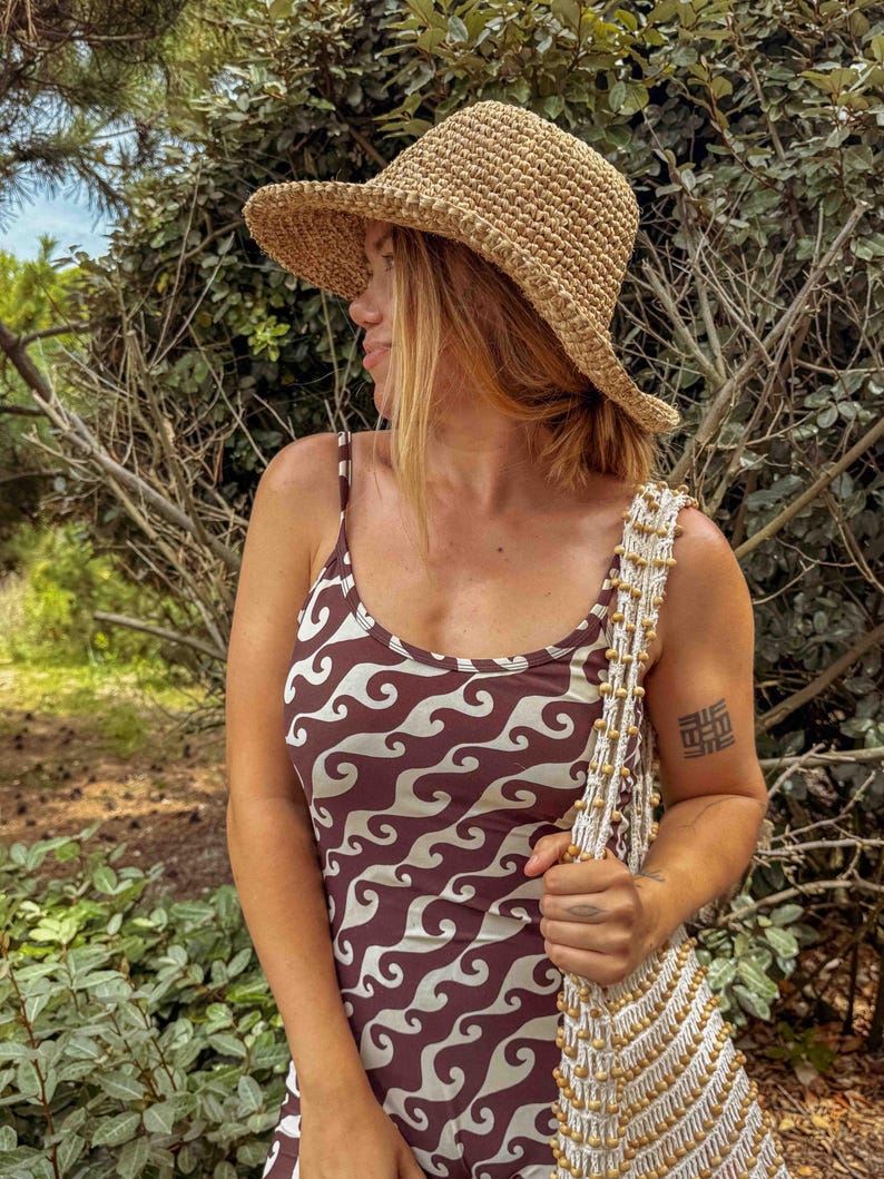 Pu&ograve; includere: Un cappello da sole in paglia intrecciata con tesa larga. &Egrave; abbinato a un abito marrone e bianco con motivo a onde e una borsa a tracolla color crema con perline. L'immagine &egrave; scattata all'aperto con uno sfondo verde naturale.