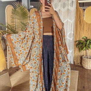 Può includere: Un lungo kimono fluente con un motivo floreale arancione, blu e bianco. Il kimono ha un ampio bordo arancione e viene indossato sopra un top a tubo marrone e pantaloni blu scuro. Lo stile generale è bohémien e rilassato.