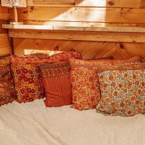 May include: Five patterned throw pillows in shades of red, orange, and blue. The pillows are arranged on a white bedspread in a wooden cabin.