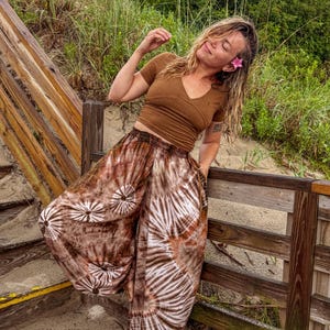 May include: A brown V-neck top and tie-dye trousers with a white and brown spiral design. The trousers have a loose, flowing fit. The outfit is worn outdoors near a wooden boardwalk and beach vegetation.