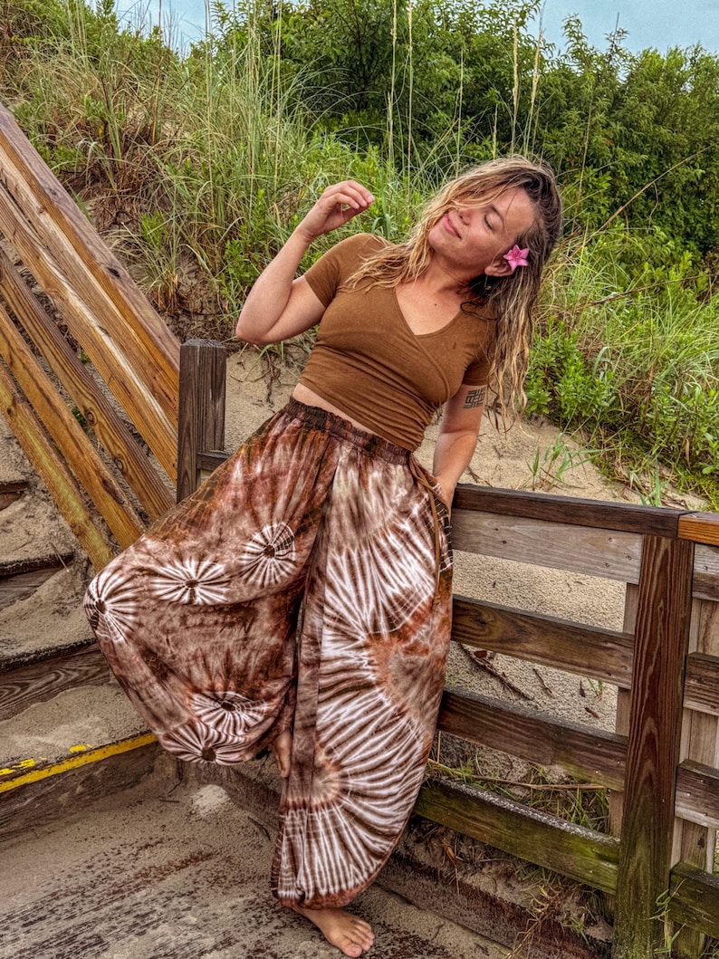May include: A brown V-neck top and tie-dye trousers with a white and brown spiral design. The trousers have a loose, flowing fit. The outfit is worn outdoors near a wooden boardwalk and beach vegetation.