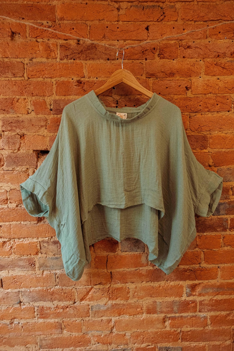 May include: A sage green, lightweight, loose-fitting, cropped top with a gathered neckline. The top has a relaxed, flowy silhouette and is made of a soft, breathable fabric.