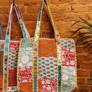 May include: Three colorful patchwork tote bags hanging on hooks against a brick wall. The bags are made of different fabrics with floral and geometric patterns in shades of orange, red, blue, green, and white.