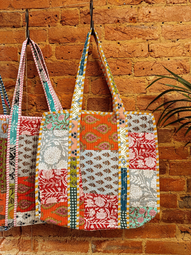 May include: Three colorful patchwork tote bags hanging on hooks against a brick wall. The bags are made of different fabrics with floral and geometric patterns in shades of orange, red, blue, green, and white.