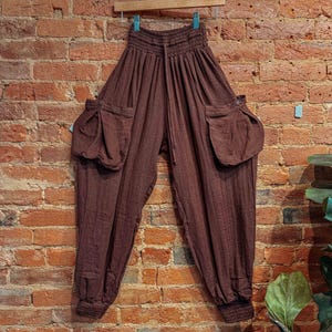 May include: Brown, loose-fitting trousers with a drawstring waist and large pockets on the sides. The trousers are hanging on a wooden hanger against a brick wall.