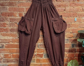 May include: Brown, loose-fitting pants with a drawstring waist and large pockets on the sides. The pants are hanging on a wooden hanger against a brick wall.