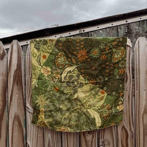 Puede incluir: Una tela acolchada en tonos verdes y oliva, adornada con motivos florales y solares, cuelga de una valla de madera desgastada. La tela presenta un dise&ntilde;o de patchwork con detalles en naranja y crema.