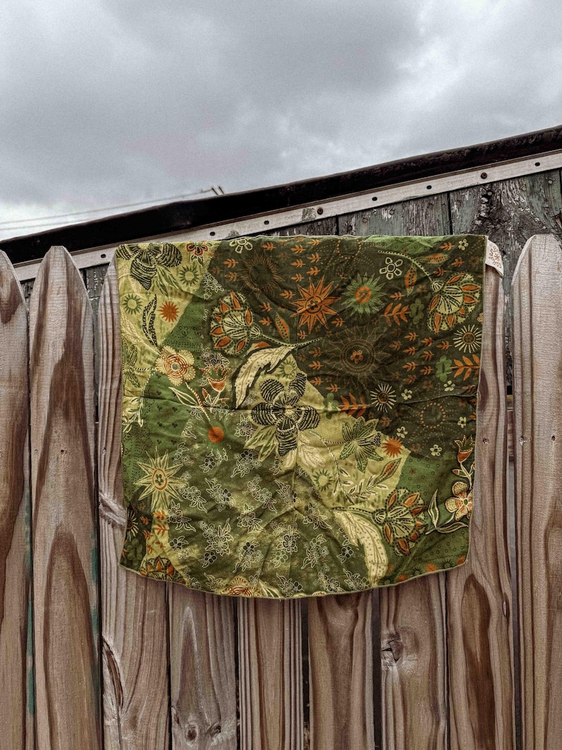 Puede incluir: Una tela acolchada en tonos verdes y oliva, adornada con motivos florales y solares, cuelga de una valla de madera desgastada. La tela presenta un dise&ntilde;o de patchwork con detalles en naranja y crema.