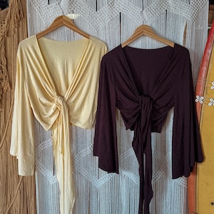 Op de afbeelding: Twee wikkel tops met lange mouwen. Eén is lichtgeel, de andere diep bordeaux. Beide tops hebben een V-hals en een striksluiting in de taille. De tops hangen aan houten kleerhangers tegen een witte macramé achtergrond.