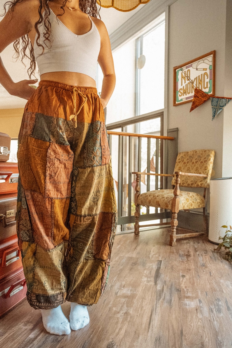 May include: A person wearing a white tank top and brown patchwork pants. The pants have a drawstring waist and are made of different fabrics. The person is standing in a room with a wooden floor and a chair.