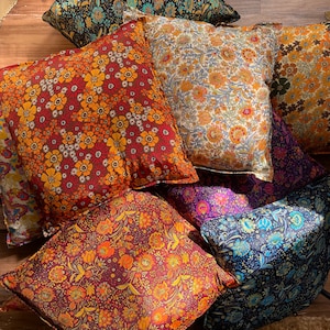 Könnte beinhalten: Eine Sammlung von sechs bunten Kissen mit Blumenmustern. Die Kissen liegen auf einem Holzboden in einem Stapel. Die Kissen sind aus einem weichen, bequemen Stoff gefertigt und in verschiedenen Farben gehalten, darunter Rot, Orange, Gelb, Blau, Grün und Lila.