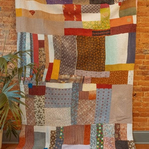 K&ouml;nnte beinhalten: Eine Patchwork-Decke aus verschiedenen Stoffen in Braun-, Rot-, Blau- und Gr&uuml;nt&ouml;nen. Die Decke besteht aus Quadraten und Rechtecken unterschiedlicher Gr&ouml;&szlig;e und Muster. Die Decke h&auml;ngt an einer Backsteinwand.