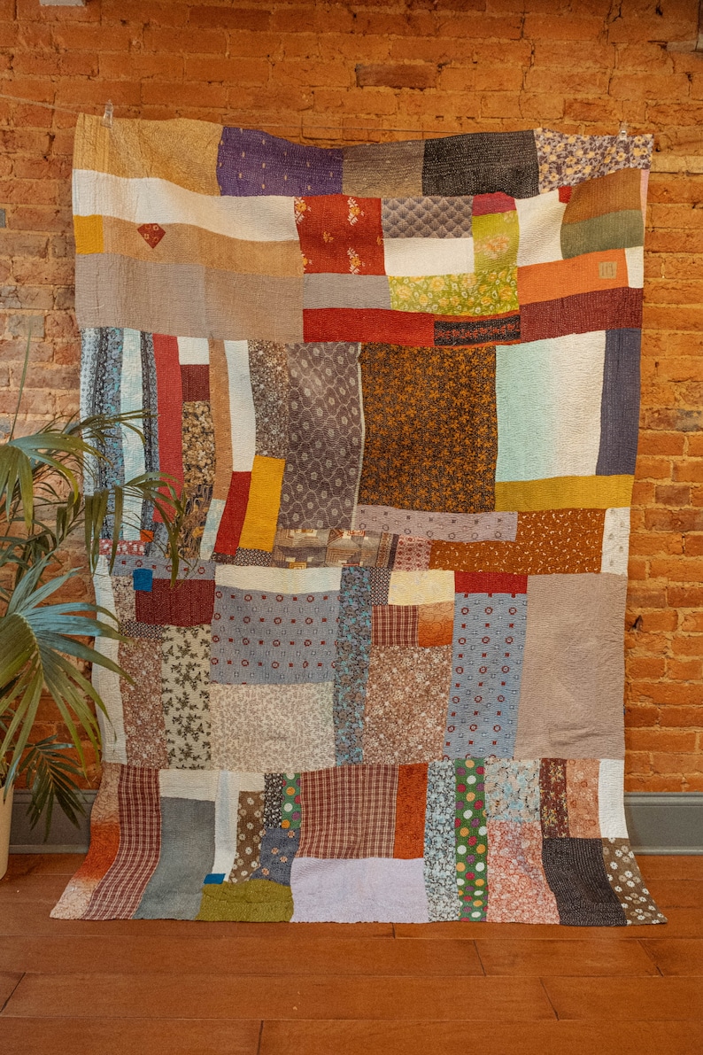 K&ouml;nnte beinhalten: Eine Patchwork-Decke aus verschiedenen Stoffen in Braun-, Rot-, Blau- und Gr&uuml;nt&ouml;nen. Die Decke besteht aus Quadraten und Rechtecken unterschiedlicher Gr&ouml;&szlig;e und Muster. Die Decke h&auml;ngt an einer Backsteinwand.