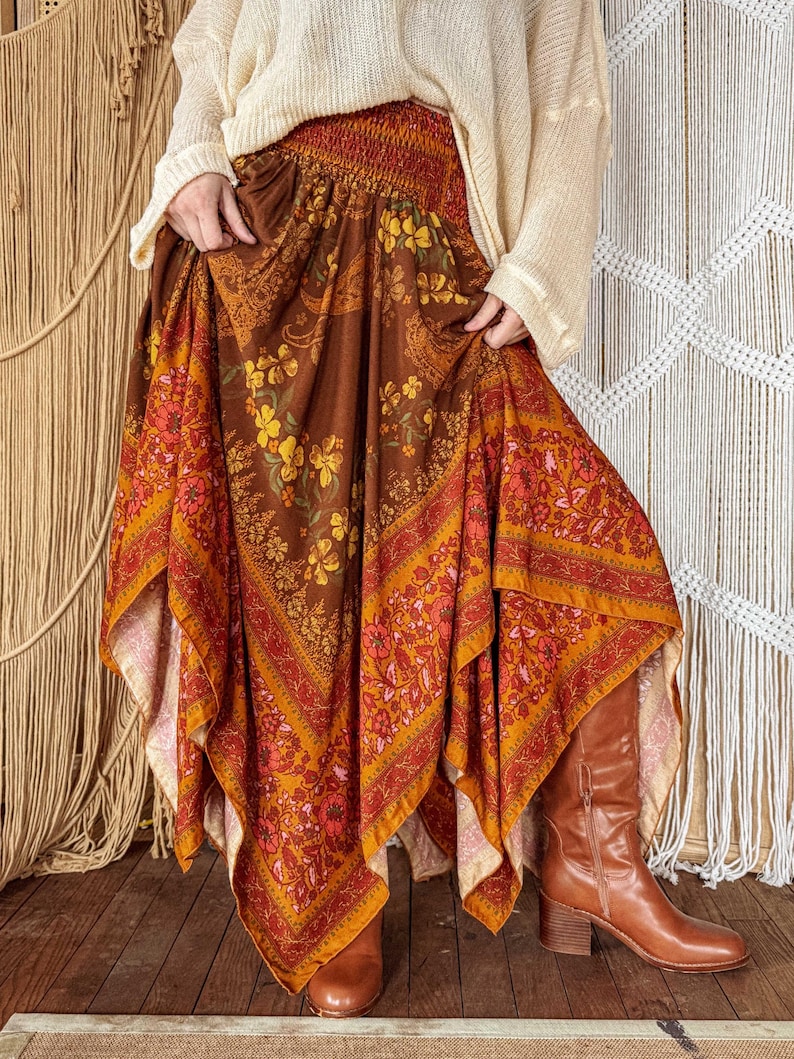 May include: A long, flowing skirt featuring a brown base with a vibrant floral and paisley pattern in shades of orange, yellow, and red. The skirt has a wide, smocked elastic waistband and an asymmetrical, handkerchief-style hemline.